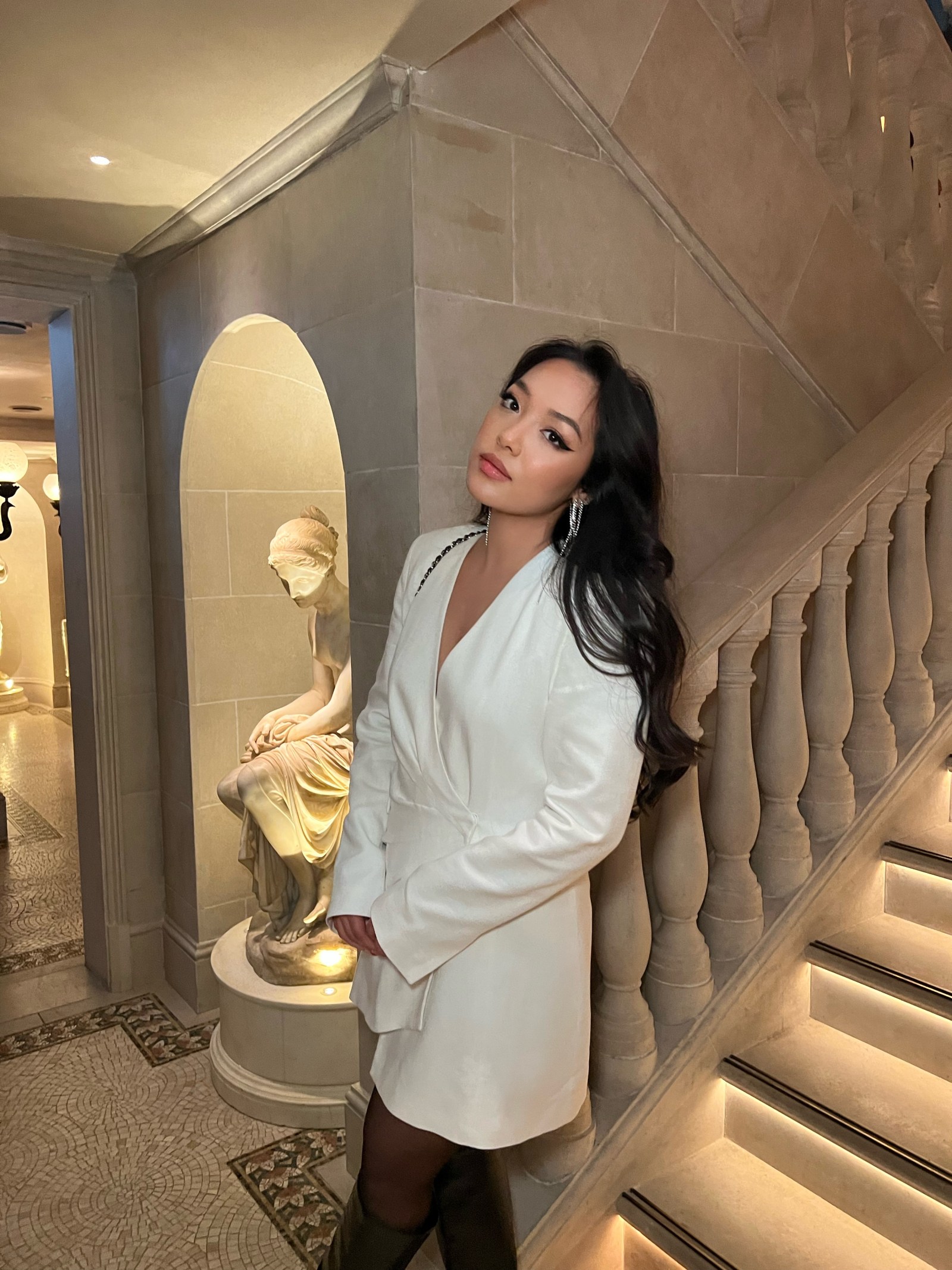 Nhi Pm in a white blazer beside a marble sculpture on a stone staircase